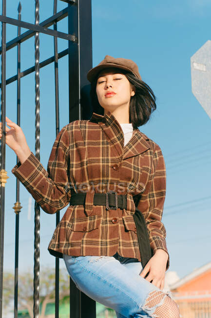 From below young ethnic female in checkered jacket with red lips looking at camera against fence in daytime — Stock Photo