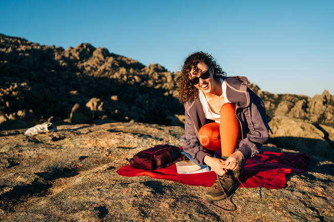Full body of positive young female traveler with long curly hair in casual clothes and sunglasses smiling while tying laces of boots on rocky mountain slope on sunny day — Stock Photo
