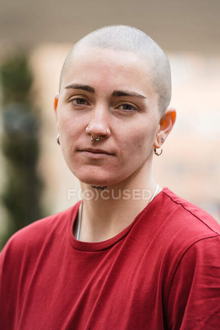 Jovem lésbica sem emoção feminina em camiseta vermelha olhando para a câmera contra a fachada da casa na cidade — Fotografia de Stock
