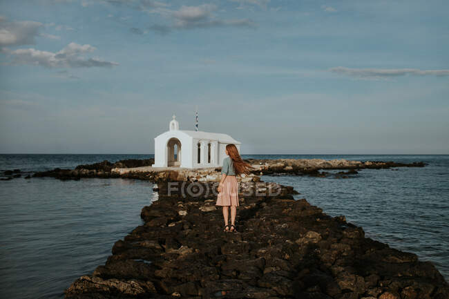 Погляд на всю довжину жінки-мандрівника, що стоїть на кам'яному острівці, що веде до білої старої церкви Святого Ніколаса під ясним блакитним небом на Криті Греції. — стокове фото