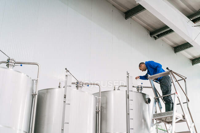 Männlicher Ingenieur auf Leiter gießt Flüssigkeit in Edelstahltank ein, während er in Brauerei arbeitet — Stockfoto