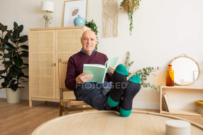 Усміхнений зрілий чоловік сидить на дерев'яному стільці і читає книгу, насолоджуючись вихідними у затишній вітальні — стокове фото