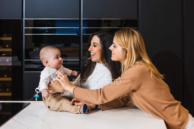 Cheerful couple of lesbian women caressing cute toddler sitting on counter in kitchen — Stock Photo