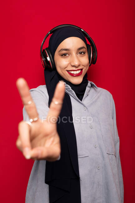 Sonriente mujer musulmana atractiva con ropa casual y hijab escuchando música favorita a través de auriculares y mostrando dos dedos sobre fondo rojo en el estudio - foto de stock