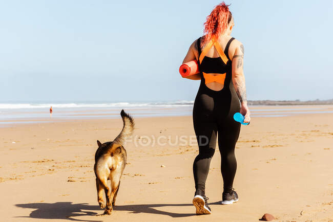 Back view of unrecognizable female athlete with rolled mat and bottle of water strolling with purebred dog on sandy sea coast — Stock Photo