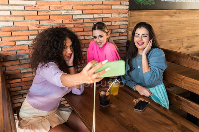 Счастливые молодые девушки в повседневной одежде делают селфи по мобильному телефону, обедая вместе в современном ресторане — стоковое фото