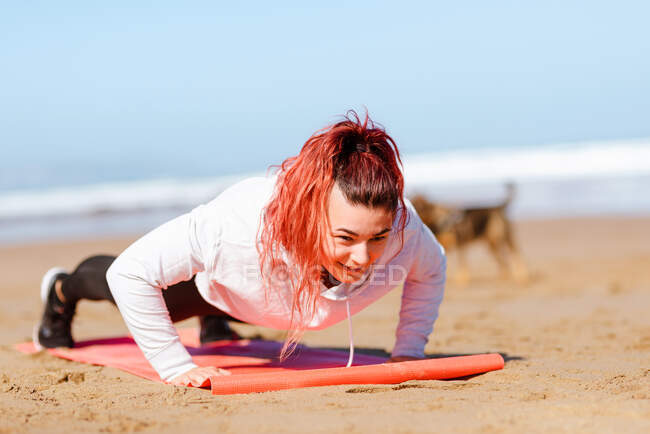 Smiling female athlete working out doing plank on mat while looking away against purebred dog on sandy coast — Stock Photo