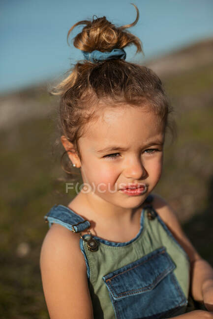 Encantada niña adorable en overoles de pie en el prado y mirando hacia otro lado - foto de stock