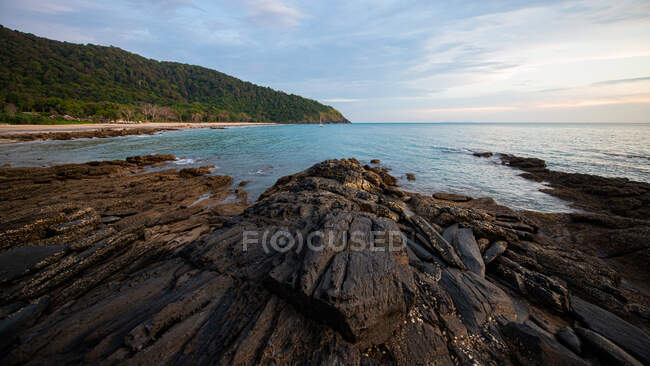 Picturesque view of rough shore against high mount and sea with horizon under cloudy sky at sundown in Thailand — Stock Photo