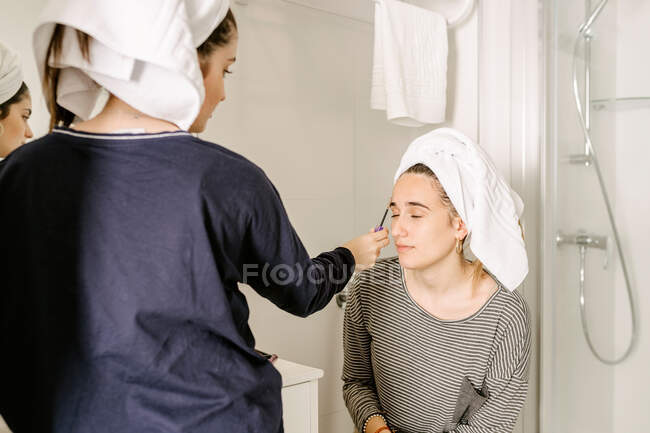 Young ethnic female in casual clothes and towel on head applying makeup on face of best friend siting in bathroom with closed eyes — Photo de stock
