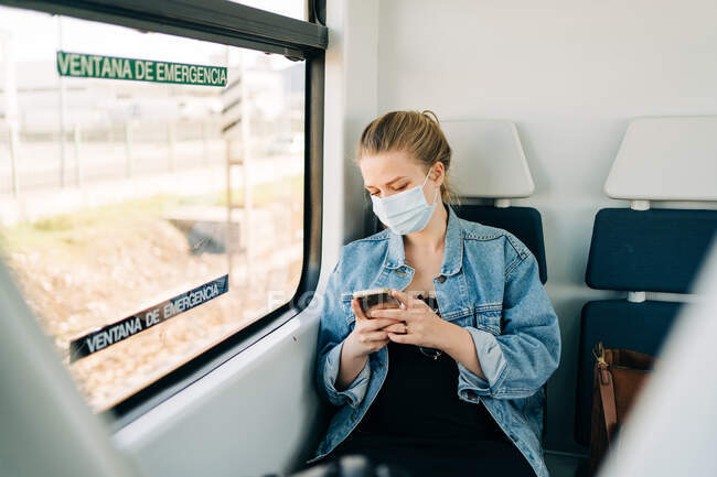 Lässige junge Frau in Jeansjacke und medizinischer Maske surft im Smartphone, während sie auf dem Zugsitz am Fenster sitzt — Stockfoto