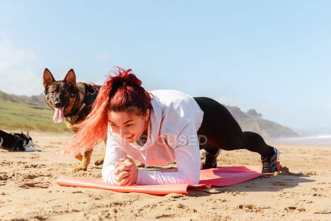 Lächelnde Athletin beim Planken auf Matte, während sie an der Sandküste auf reinrassige Hunde herabblickt — Stockfoto