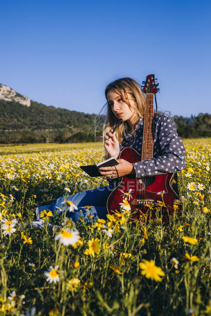 Задумчивая молодая хипстерша, сидящая на лугу в сельской местности, пишет песни на блокноте и играет на гитаре во время летнего солнечного света, отводя взгляд — стоковое фото