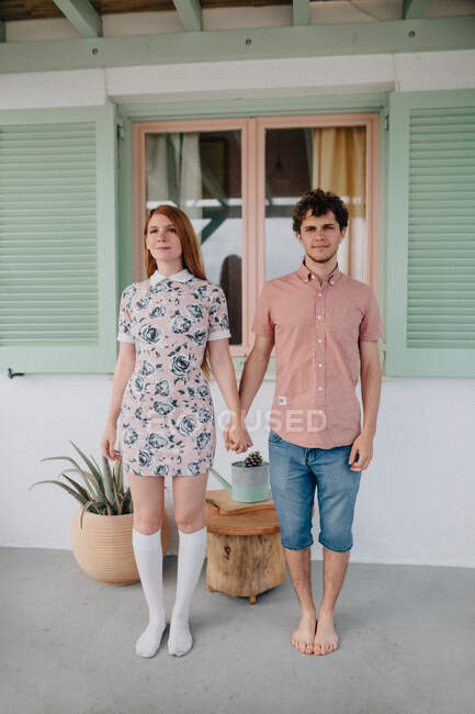 Полное содержание тела молодая пара носить случайный летний наряд держаться за руки и смотреть на камеру, стоя рядом с современным небольшим коттеджем — стоковое фото