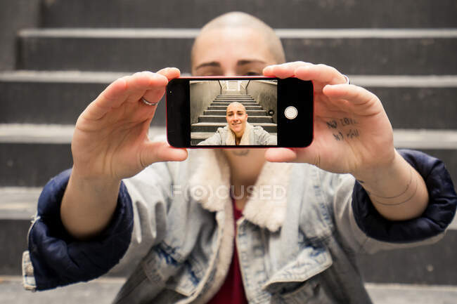 Весела транссексуальна людина з татуюванням знімає власний портрет на мобільному телефоні, сидячи на сходах і дивлячись на камеру — стокове фото