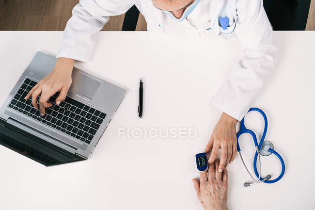Топ-представление о неузнаваемом враче, измеряющем уровень кислорода в крови пациента с монитором сердечного ритма за столом со стетоскопом в клинике — стоковое фото