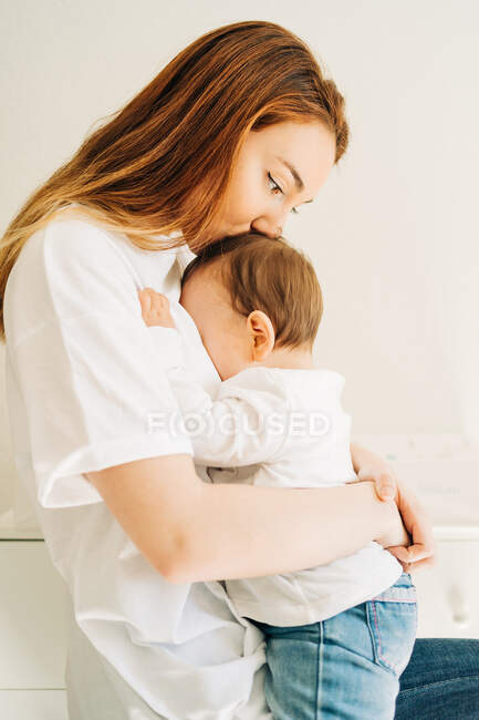 Seitenansicht junge Mutter in lässiger Kleidung umarmt und küsst kleines Baby im hellen Raum — Stockfoto