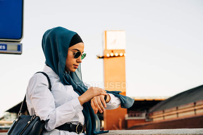 Vista lateral de la mujer musulmana en hijab de pie en la plataforma en la estación de tren y comprobar la hora en el reloj de pulsera mientras espera el tren - foto de stock
