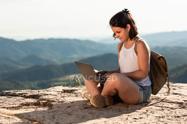 Vista lateral del explorador femenino sentado en la roca en las montañas y netbook de navegación durante las vacaciones de verano - foto de stock