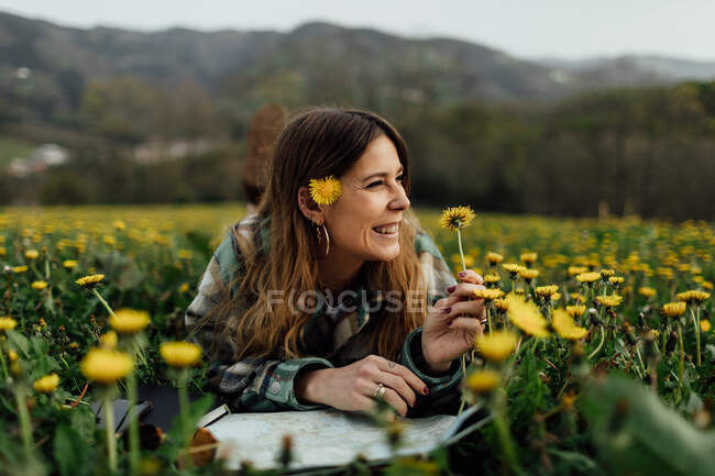 Content female traveler with paper map and blossoming flowers looking away while lying on meadow against mountain in countryside - foto de stock