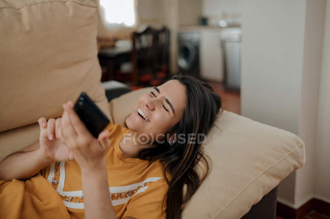Junge Frau schreibt SMS auf Handy, während sie sich auf Couch im Wohnzimmer hinlegt — Stockfoto