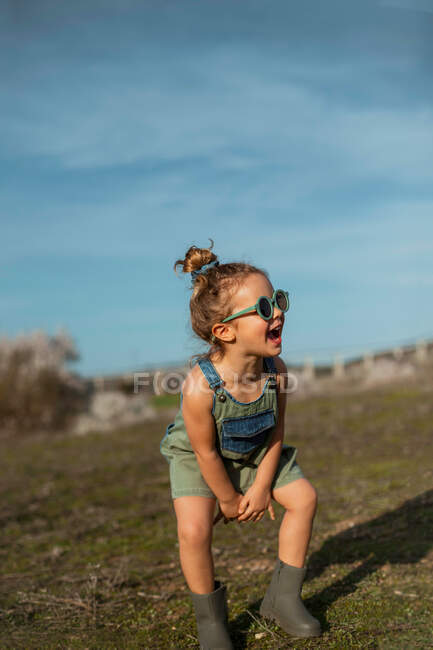 Content little girl in overalls and sunglasses jumping with outstretched arms above meadow and enjoying summer on sunny day in countryside — Stock Photo