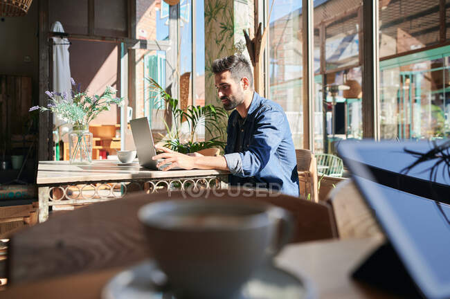 Bearded ethnic male remote employee typing on netbook at table with cup of coffee in sunlight — Stock Photo