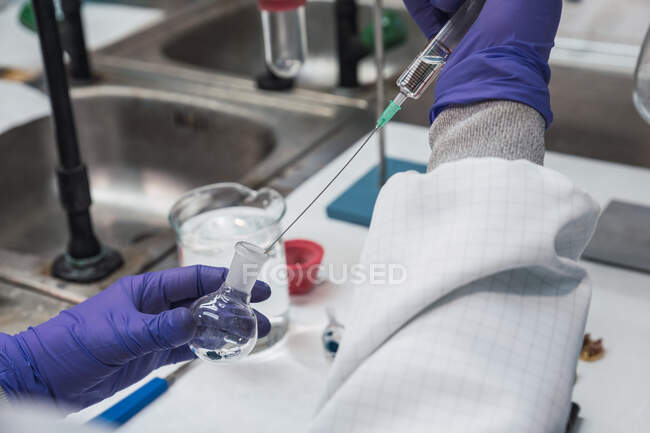 Crop unrecognizable scientist in white robe and gloves conducting chemical experiment with substance and syringe while working in modern laboratory — Stock Photo