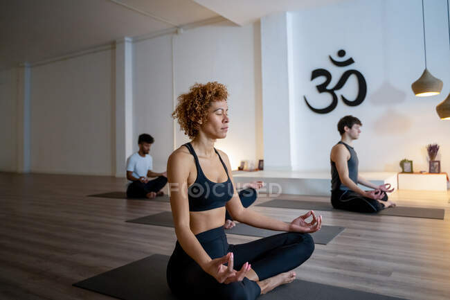 Mulher afro-americana com grupo de diversas pessoas sentadas em Lotus posar com os olhos fechados e mediar enquanto praticam ioga juntos durante a aula em estúdio — Fotografia de Stock