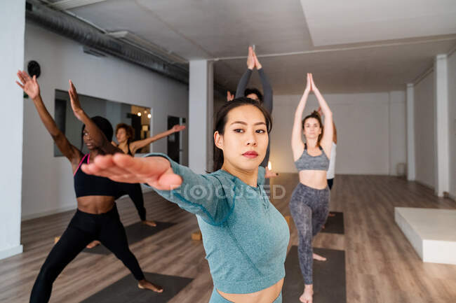 Focalizzato femminile asiatica in piedi in Virabhadrasana e pratica consapevolezza durante la lezione di yoga con diverse persone in studio — Foto stock