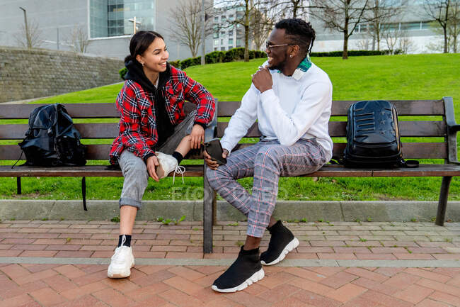 Feliz casal multiétnico em roupas elegantes falando enquanto olham um para o outro no banco da cidade à luz do dia — Fotografia de Stock