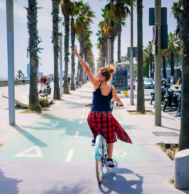 Back view of cool female bicyclist riding bike while demonstrating victory gesture with raised arm on city walkway - foto de stock