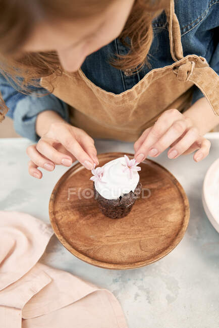 Обрізати молоду жінку, що прикрашає шоколадний кекс з солодким збитими вершками та квітами на підставці для торта, готуючи вдома — стокове фото