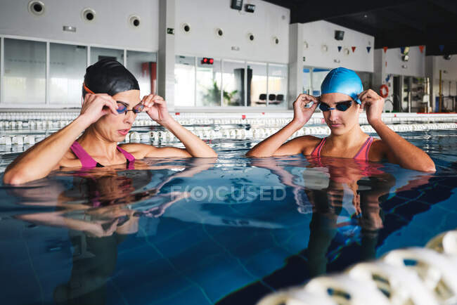 Femmes sportives en bonnets et maillots de bain se préparant pour l'entraînement dans la piscine avec de l'eau transparente pendant la journée — Photo de stock