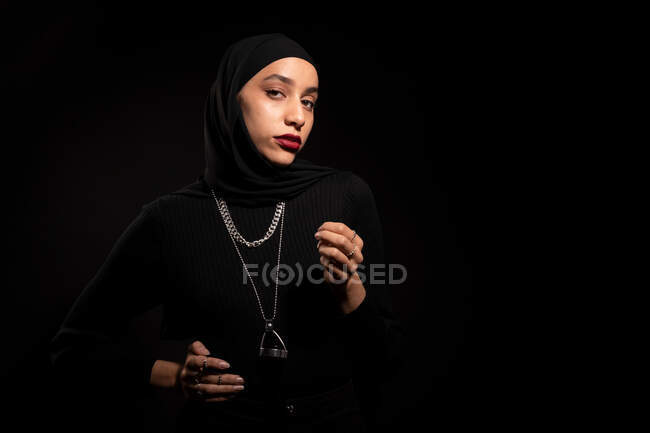 Attractive young Islamic female wearing black outfit and hijab gently looking at camera on black studio — Stock Photo