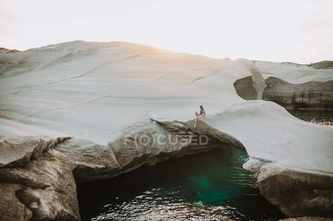 Distant fit female in swimsuit chilling on uneven stony seashore washed by azure rippling sea in sunny Sarakiniko Milos — Stock Photo