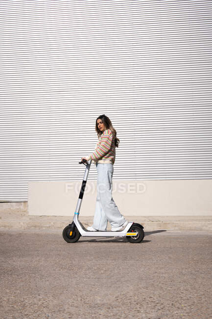 Вид сбоку на молодую женщину в стильном наряде, катающуюся на электроскутере по асфальтовой дороге в городе — стоковое фото