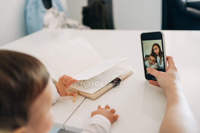 Обрізати молоду маму і чарівну дитину взявши селфі на сучасному мобільному телефоні, сидячи за столом разом — стокове фото