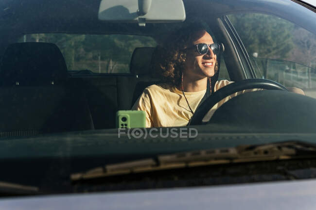 Happy young haired male in stylish sunglasses looking away through open window of car while sitting at driver seat — Photo de stock