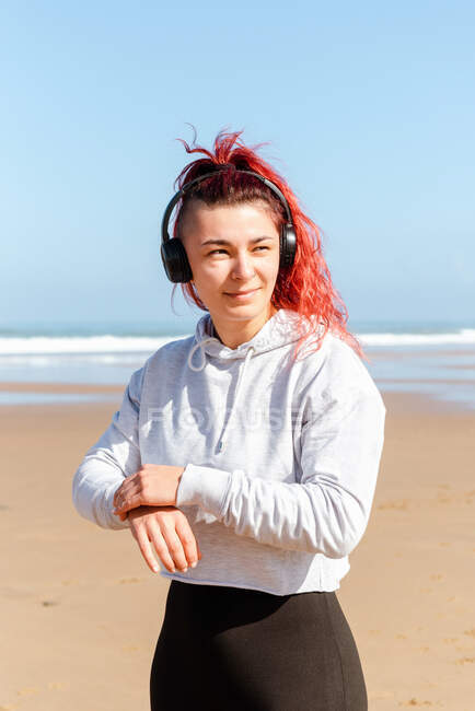 Smiling female athlete in sports clothes and headphones looking away on ocean beach during break from workout — Stock Photo