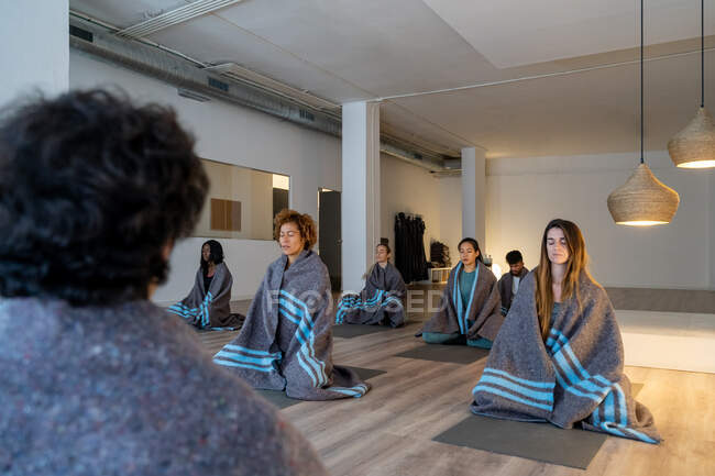 Personas tranquilas multirraciales envueltas en cuadros sentados en esteras en estudio y meditando durante la clase de yoga - foto de stock
