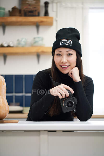 Contenu femme ethnique avec appareil photo numérique penché avec la main sur la table tout en regardant l'appareil photo — Photo de stock