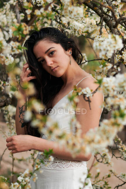 Young female with tattooed arm wearing white dress and standing in flowers of tree looking at camera — Foto stock