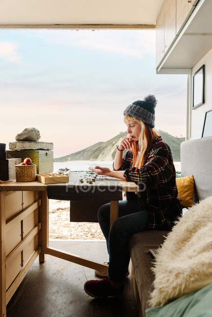 Side view of focused female traveler creating handmade accessories while sitting at wooden table in parked truck at seaside — Stock Photo
