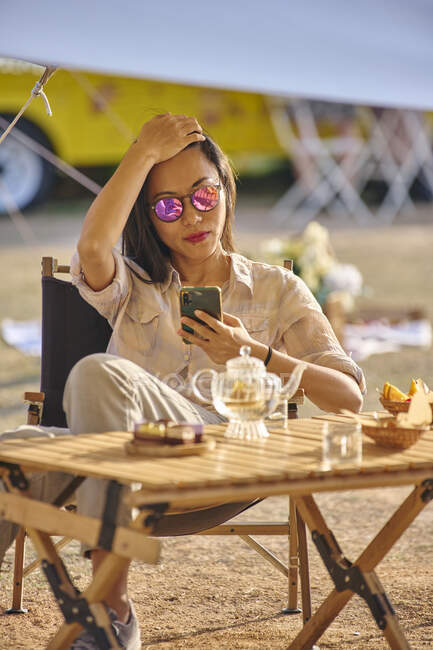 Hermosa chica asiática navegando en el teléfono móvil, mientras que tener un tiempo relajante sentado en la mesa en el área de camping - foto de stock