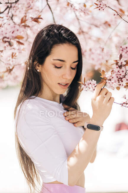 Young alluring female with curved body looking down under almond blossom tree on blurred background — Foto stock