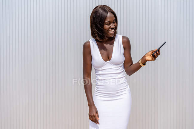 Mujer afroamericana con estilo positivo en vestido blanco que navega por el teléfono móvil moderno mientras está de pie cerca de la pared del edificio en la calle soleada - foto de stock