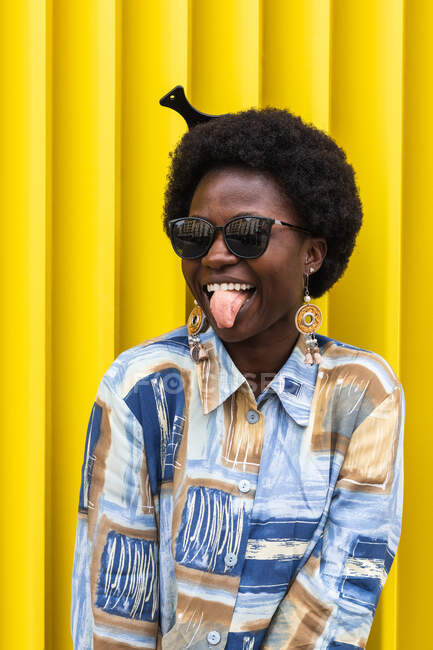 Игривая молодая африканская американка с афроволосами в стильной красочной рубашке в солнечных очках и сережках, развлекающаяся и показывающая язык у желтой стены — стоковое фото