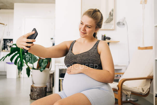 Junge zufriedene werdende Frau sitzt auf Fitnessball, während sie zu Hause ein Selfie mit dem Handy macht — Stockfoto