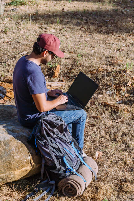 Visão lateral corpo inteiro de nômade sentado em pedra perto de pertences e usando netbook enquanto trabalhava remotamente — Fotografia de Stock